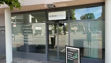 The image shows the exterior of a modern building with large glass windows and a sign reading "AURUM HEALTH & PERFORMANCE". A promotional sign in front advertises a training program. The building has a clean, contemporary design with a visible entrance an