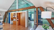 Bright, modern room with wooden flooring, large glass windows, and exposed wooden beams. Features a white table with a potted plant, chairs, a wooden cabinet with shelves, and a desk area with a telephone and books.