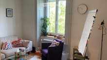 Living room with a sofa, armchair, coffee table, clock, plant, board, lamp, and picture frame. The room has large windows and a view of greenery outside.