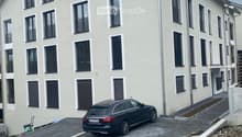 Modern two-story residential building with light beige facade, multiple windows, and balconies. A black car is parked in the driveway.