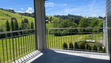 Balcony with metal railing, tiled floor, overlooking green hills and trees under a clear blue sky.
