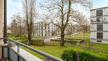 Balcony overlooking a landscaped garden area with trees, modern apartment buildings, and a road with parked cars.