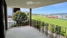 Spacious balcony with tiled floor, metal railing, and scenic view of a green field and town.