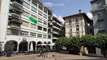 Multi-story building with balconies and green shutters, located in an urban area with outdoor seating, a fountain, and trees.