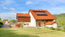 Two-story house with red-tiled roof, solar panels, stone and wood exterior, surrounded by lawn and trees.
