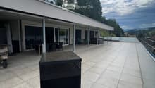 Modern outdoor patio with tiled flooring, glass railing, and a covered seating area with tables and chairs. A black barbecue grill is placed on the patio.
