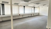 Empty room with grey carpet, white walls, large windows with blinds, and a perforated white ceiling with recessed lighting.