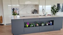 Modern kitchen with a grey island, white cabinets, marble backsplash, and wooden flooring. Features a coffee maker, sink, and decorative plants on the island and shelves.