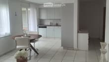 Bright, modern room with tiled flooring, a round wooden dining table with four white chairs, a vase with white flowers on a small table, and a kitchen area with white cabinets and a sink.
