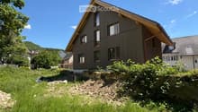 Modern two-story house with wooden siding, multiple windows, and a sloped roof, situated on a raised ground floor with surrounding greenery and a clear blue sky.