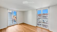 Empty room with light wooden flooring and white walls, featuring large sliding glass doors that open to a balcony with a view of rooftops.