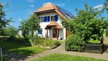 Two-story house with white walls, blue shutters, red-tiled roof, and solar panels on the roof. The house has a small porch with a wooden door and is surrounded by a well-maintained garden with a gravel pathway and wooden fence.