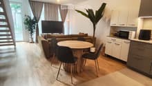 Open-plan living and dining area with a sofa, dining table, chairs, and a television. Kitchen area features white cabinets and a microwave. Wooden flooring throughout.