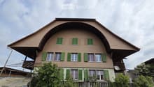 Two-story house with brown tiled roof and green shutters, surrounded by trees and a small garden area.