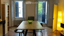 Dining area with wooden herringbone floor, modern table with four stools, green bowl on table, large windows with shutters, warm lighting from pendant lamp and floor lamp.