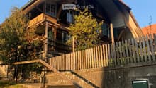 Two-story wooden house with a sloped roof, multiple windows, and a wooden balcony, surrounded by a wooden fence and greenery.