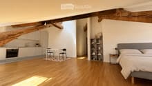 Open-plan living space with a bedroom and kitchen area. Features a wooden floor, exposed wooden beams, a grey upholstered bed, a nightstand, a small table with chairs, a kitchen with white cabinets, an oven, and a shelf with decorative items.
