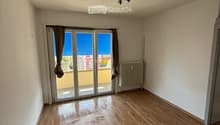 Empty room with wooden flooring, white walls, and a large sliding glass door leading to a balcony. The room has a ceiling light fixture and a radiator on one wall.