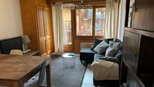 A cozy living room with wooden furniture, a sofa, coffee table, and a dining table. Large windows offer a view of the balcony and surrounding area.
