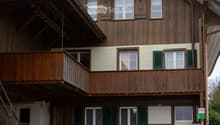 Two-story wooden house with green shutters, a wooden balcony, and a small porch area with a bench and potted plants.