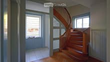 Spacious hallway with wooden flooring, a wooden staircase leading upwards, and large windows providing natural light. The walls are painted in neutral tones, and there is a small window seat with a few decorative items on the left side.