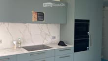 Modern kitchen with light blue cabinets, marble backsplash, induction stove, and double oven. Countertop has a few items including a bottle and a hat.