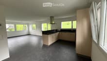 Empty kitchen with light wooden cabinets, black countertops, and large windows overlooking a green landscape.