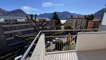 Balcony with metal railing overlooking a scenic view of mountains, a lake, and surrounding buildings. Several parked cars are visible below.