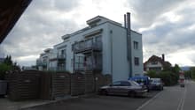 Modern multi-story building with balconies, surrounded by a wooden fence and parked cars.