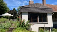 Single-story house with a modern design, featuring a raised ground floor, large windows, and a well-maintained garden with colorful flowers and an umbrella.