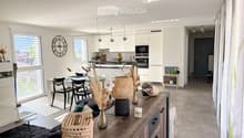 Open-plan living and dining area with modern furniture, large windows, and a kitchen area in the background.