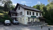Two-story building with a sign reading "TAVERNE" and "BEIZLI" on the facade, surrounded by greenery and parked cars.