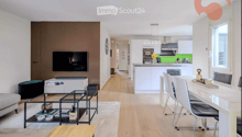 Modern open-plan living and dining area with a sofa, coffee table, TV, and a dining table with chairs. The kitchen features white cabinets, a kitchen island, and various appliances.
