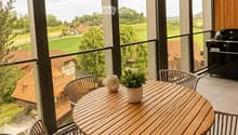 Balcony with a round wooden table, four white wicker chairs, a small potted plant, and a decorative vase. Large windows offer a scenic view of a countryside landscape.