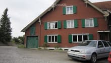 Two-story red brick house with green shutters, multiple windows, and a gravel driveway with a parked car.