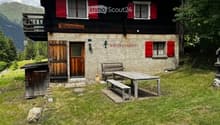 Traditional wooden chalet with red shutters, stone walls, and a balcony overlooking a mountain view. Outdoor seating area with a picnic table and benches.