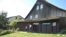 A two-story wooden house with a steep, dark wooden roof and multiple windows. The house has a covered entrance area with large windows and a door. The surrounding area includes a grassy lawn and some trees.