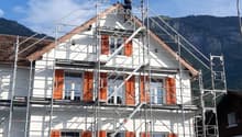 A two-story house under construction with scaffolding, orange shutters, and a worker on the roof. A truck is parked nearby.