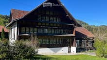 Traditional two-story wooden house with a steeply pitched roof, multiple windows, and a covered balcony area. The house features a mix of white and dark wood siding, with a well-maintained lawn and surrounding greenery.