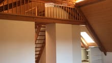 An attic space with wooden beams, exposed wooden ceiling, wooden floor, and a staircase leading to an upper level. The room is empty with white walls and a small window letting in natural light.