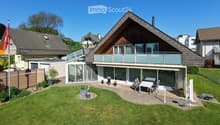 A modern two-story house with a triangular roof, large windows, and a spacious balcony. The property features a well-maintained lawn, patio area with outdoor furniture, and surrounding greenery.