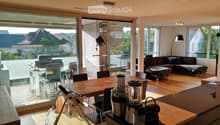 Open-plan living and dining area with wooden flooring, large windows, a sofa, a coffee table, a dining table with chairs, and a kitchen counter with a coffee machine and blender.