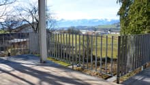 Balcony with metal railing, tiled floor, overlooking a green field and distant mountains.