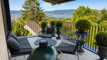 Balcony with wooden decking, black metal railing, and scenic mountain and lake view. Features a green grill, sofa, chairs, and potted plants.