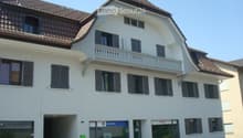 Building. A two-story white building with a brown tiled roof, multiple windows with shutters, and a small balcony on the upper floor. The ground floor features a glass entrance with a sign that reads "ZURICH".
