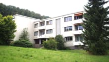 Modern multi-story residential building with balconies, surrounded by greenery and trees.