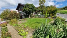 A well-maintained garden area with a variety of plants, shrubs, and flowers, leading up to a traditional Swiss house with a sloped roof and green shutters.