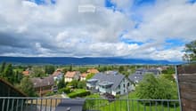 Balcony overlooking a suburban neighborhood with houses, green fields, and distant mountains under a partly cloudy sky. A grill is placed on the balcony.