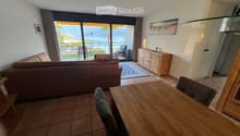 Bright living room with a view of the sea, featuring a sofa, dining table, chairs, and a cabinet.