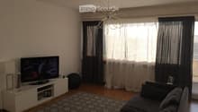 Modern living room with a grey sectional sofa, a flat-screen TV on a white stand, a patterned rug, and large windows with dark curtains. A decorative lamp hangs from the ceiling.